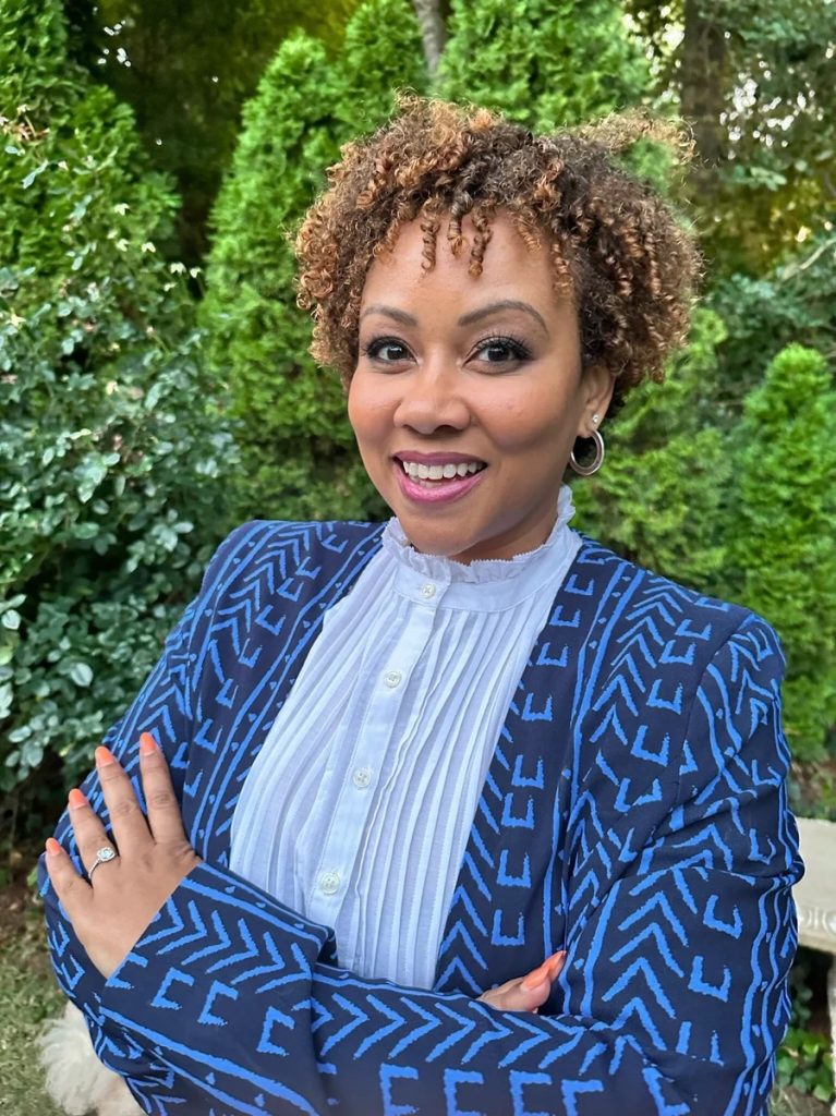 Shaunté outside with arms folded, wearing blue in front of foliage